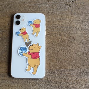 May include: Three Winnie the Pooh phone stickers. Each sticker features Winnie the Pooh holding a jar of honey that says "SMUT" on it. The stickers are on a white iPhone.