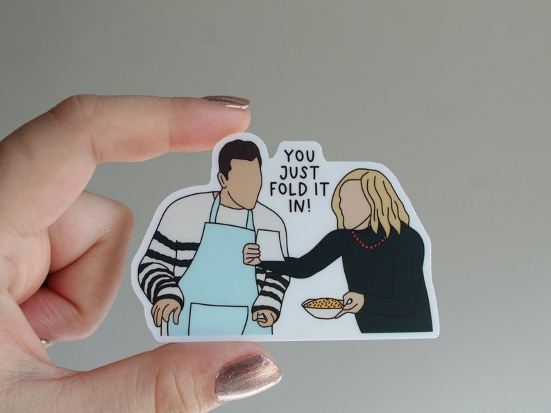 May include: A sticker featuring a cartoon illustration of two people, one wearing a blue apron and the other wearing a black shirt and a red necklace. The text "You just fold it in!" is written in white on a black background.