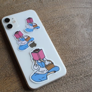 May include: Three phone case stickers featuring a cartoon woman reading a book. The woman is wearing a blue dress and a brown basket. The stickers are on a white phone case.