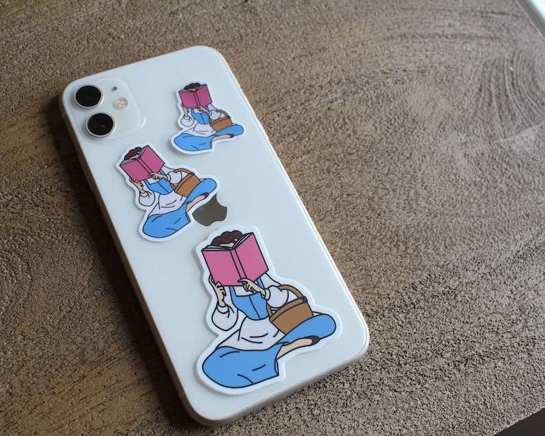 May include: Three phone case stickers featuring a cartoon woman reading a book. The woman is wearing a blue dress and a brown basket. The stickers are on a white phone case.