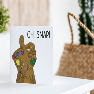 May include: A white card with a gold hand wearing a gauntlet with colorful gems. The text "OH. SNAP!" is printed in black on the card.