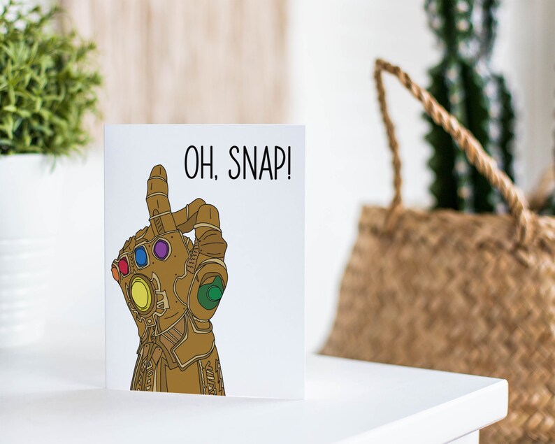 May include: A white card with a gold hand wearing a gauntlet with colorful gems. The text "OH. SNAP!" is printed in black on the card.