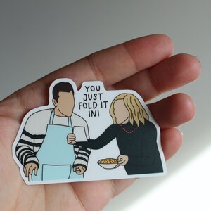 May include: A sticker featuring a cartoon illustration of two people, one holding a bowl of cereal and the other holding a mug. The text on the sticker reads "You just fold it in!"