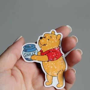 May include: A Winnie the Pooh sticker with glitter accents. Pooh is holding a blue jar that says "SMUT" on it.