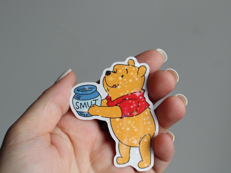 May include: A Winnie the Pooh sticker with glitter accents. Pooh is holding a blue jar that says "SMUT" on it.