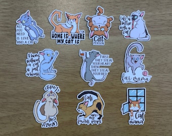 May include: A collection of cat-themed stickers with various designs and text. The stickers feature cartoon cats in different poses, with phrases like "All you need is love and a cat" and "Home is where my cat is."