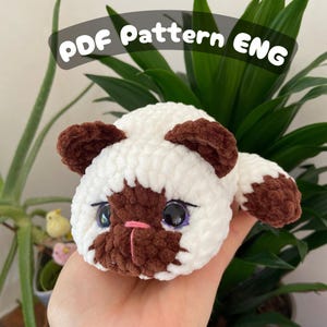Puede incluir: Un juguete de gato hecho a mano, de color blanco y marrón. El gato tiene ojos grandes y negros con detalles morados, una nariz rosa y orejas y patas marrones. El texto "PDF Pattern ENG" está en la parte superior.