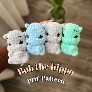 Peut inclure: Quatre petits hippopotames crochetés, dans les tons de gris, de bleu et de vert. Chaque hippopotame a des yeux noirs et une expression légèrement triste. Le texte "Bob the hippo PDF Pattern" est affiché.