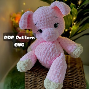 May include: A handmade pink crochet pig plush toy with white accents on its feet and snout. The pig has black eyes and is sitting on a woven basket. The text "PDF Pattern ENG" is displayed on the image.