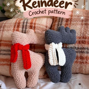 May include: Two plush reindeer toys made of soft yarn. One reindeer is light pink with a bright red scarf, and the other is dark gray with a white scarf. Above them, a graphic overlay displays the text Reindeer Crochet pattern.