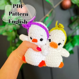 Peut inclure: Deux peluches de canard blanches au crochet avec des becs orange et des yeux noirs. Un canard porte un bandeau violet et l'autre un bandeau vert.  PDF Pattern English.
