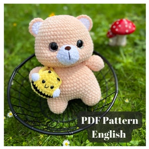 Peut inclure: Un ours en peluche rose en crochet tenant un jouet d'abeille en crochet jaune et noir. L'ours a des yeux noirs avec des paillettes, un nez blanc et une bouche blanche. L'abeille a un visage souriant. Les jouets sont assis dans un panier en fil de fer noir sur un lit d'herbe verte. Le texte "PDF Pattern English" est en bas de l'image.