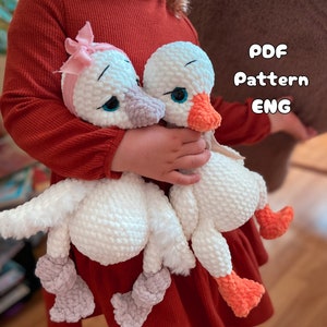 Peut inclure: Deux jouets amigurumi en forme de cygne en crochet blanc avec des accents roses et orange. Les jouets sont tenus par un enfant portant un pull rouge. L'image comprend le texte "PDF Pattern ENG".