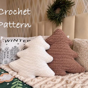 Può includere: Due cuscini all'uncinetto a forma di albero in color crema e marrone, posizionati davanti a un cuscino rettangolare bianco con un disegno a linee nere di edifici e il testo WINTER, accanto a un cuscino beige testurizzato e una coperta verde.