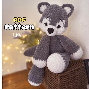 May include: A handmade grey and white crocheted wolf plush toy. The wolf has a black nose, white accents around the eyes and mouth, and white paws. The text "PDF Pattern ENG" is in the upper left corner. The plush is sitting on a woven basket.