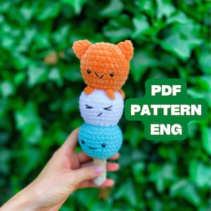 Może przedstawiać: Ręcznie robiona zabawka w kształcie rożka lodowego z szydełka z trzema kolorowymi gałkami: pomarańczową, białą i niebieską. Każda gałka ma uśmiechniętą buzię. Zabawka jest trzymana na tle zielonego, liściastego tła. Tekst na obrazku brzmi "PDF PATTERN ENG".