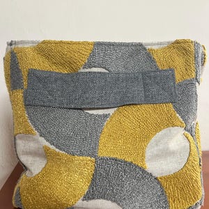 May include: A rectangular clutch with a textured surface. The design features a pattern of yellow, gray, and off-white shapes. A gray fabric band is sewn across the front. The clutch is made of a soft, woven material.