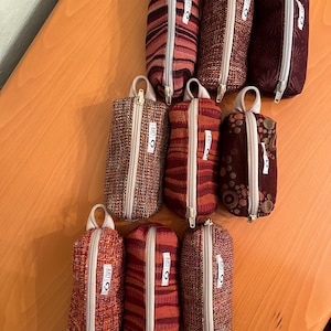 May include: A collection of small, cylindrical fabric pouches in various shades of red, brown, and burgundy. Each pouch features a white zipper, a small loop handle, and a tag with the word "LIFE". The pouches have different patterns and textures.