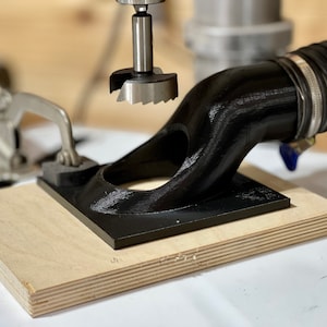 May include: A black 3D printed dust collection attachment for a drill press. The attachment is mounted on a piece of plywood and is connected to a black hose. A drill bit is visible in the background.