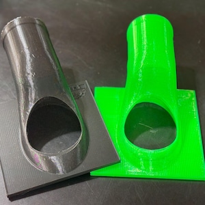May include: Two 3D printed plastic nozzles, one black and one green, with a circular opening. The nozzles are mounted on square bases.