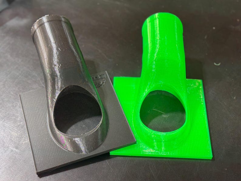 May include: Two 3D printed plastic nozzles, one black and one green, with a circular opening. The nozzles are mounted on square bases.