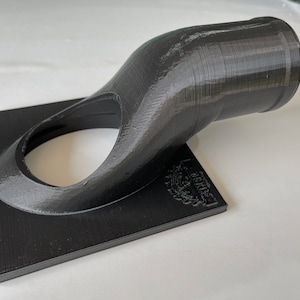 May include: A black 3D printed object with a curved spout and a square base. The object has a small hole in the centre of the base and the text "Le Creuset" is printed on the base.