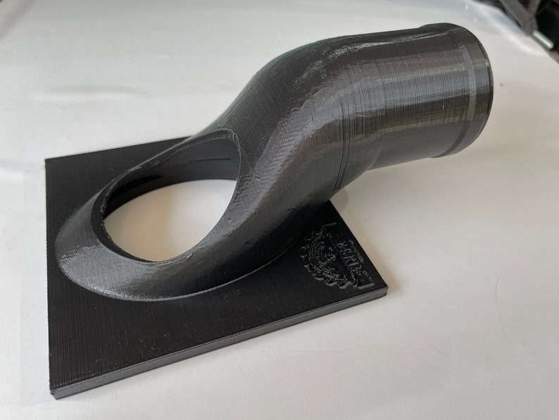 May include: A black 3D printed object with a curved spout and a square base. The object has a small hole in the centre of the base and the text "Le Creuset" is printed on the base.