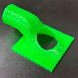 May include: A bright green 3D printed plastic object with a curved tube and a square base. The tube has a round opening on one end and a smaller round opening on the other end.