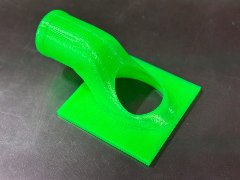 May include: A bright green 3D printed plastic object with a curved tube and a square base. The tube has a round opening on one end and a smaller round opening on the other end.