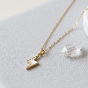 May include: A gold-colored necklace with a lightning bolt pendant. The pendant is white with gold trim. A clear crystal and a white ceramic object are next to the necklace on a gray surface. The necklace chain is delicate.