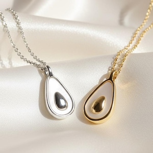 May include: Two avocado-shaped pendant necklaces. One is silver, the other gold. Each pendant has a white interior and a dark brown seed. The necklaces are on delicate chains, displayed on a white, silky surface.