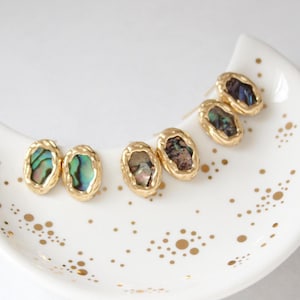 Puede incluir: Pendientes de botón dorados con un marco ovalado texturizado. Los pendientes presentan inserciones de concha de abulón iridiscente en tonos verdes, azules y morados. Los pendientes se exhiben en un plato blanco con motas doradas.