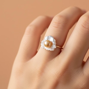 May include: A gold ring with a white, petal-shaped setting holding a golden pearl. The ring is displayed on a finger, against a neutral backdrop. The design of the ring and the warm colours create an elegant aesthetic.