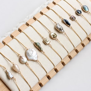 May include: A collection of delicate gold-tone chain bracelets, each featuring a single irregular baroque pearl in various shades including white, cream, grey, green, and dark blue, arranged in a row on a light-coloured display stand.