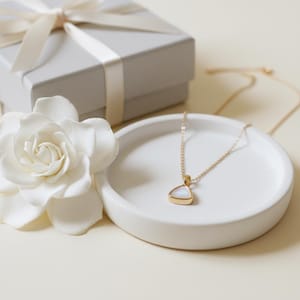 May include: A gold necklace with a triangular pendant containing a white stone rests on a white ceramic dish. A white flower and a grey gift box with a white ribbon are also in the image. The jewellery is likely a necklace.