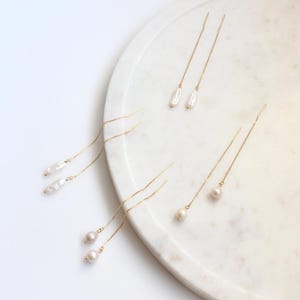 Peut inclure: Quatre paires de boucles d'oreilles pendantes en or avec des accents de perles. Les perles sont blanches et varient en forme et en taille. Les boucles d'oreilles sont présentées sur une surface en marbre blanc.