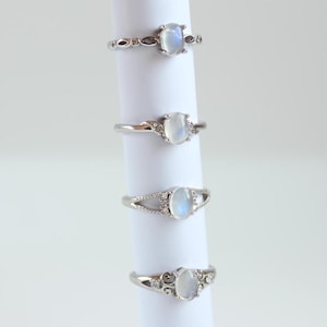 May include: Four silver rings with oval moonstone settings. The rings have different styles of bands, including a simple band, a band with small stones, a band with a delicate openwork design, and a band with a floral design.