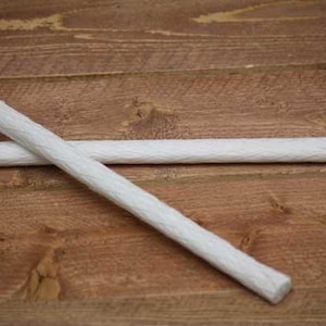 May include: Two white cylindrical candle wicks with a braided texture. The wicks are long, thin, and straight, positioned in a crossed arrangement. The material appears to be a woven fiber, showing a consistent pattern along the entire length.