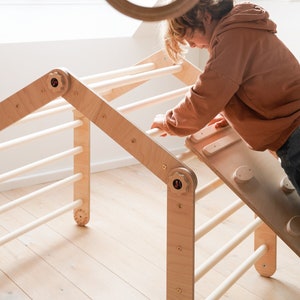 May include: A child plays on a wooden climbing frame with a slide and a ladder. The frame is made of natural wood and has a light brown finish. The child is wearing a brown hoodie and is reaching up to grab a wooden bar.
