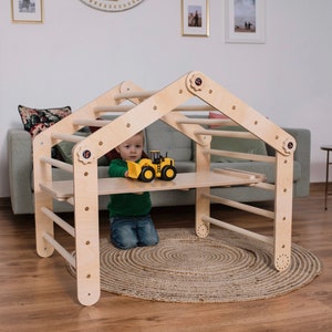 May include: A wooden climbing frame shaped like a house with a child playing inside. The frame has a ladder, a slide, and a platform. The child is holding a toy truck.