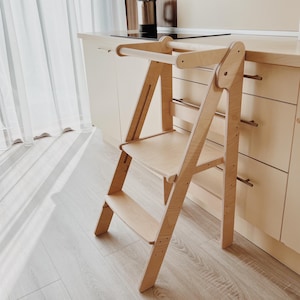 May include: A light wood children's step stool with a safety rail and a step. The stool is in a kitchen setting.