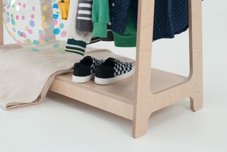 Op de afbeelding: Een houten kledingrek met een plank voor schoenen. Het rek is gemaakt van licht hout en heeft een eenvoudig ontwerp. Er staan twee paar schoenen op de plank, een paar is zwart met een wit geruit patroon en het andere paar is zwart.