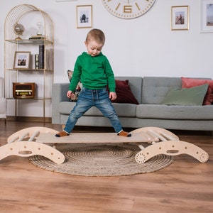 May include: A child in a green hoodie and blue jeans stands on a wooden balance beam with a ladder on one side. The beam is curved at both ends and has a handle in the center. The beam is on a light brown wood floor with a woven rug underneath.