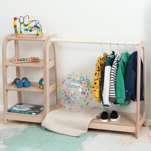 Op de afbeelding: Een houten kledingrek voor kinderen met een rek. Het rek heeft een hangende stang met kleding op hangers en een lager rek. Het rek heeft drie planken met speelgoed erop. Er ligt een kleurrijke bal op de vloer voor het rek.