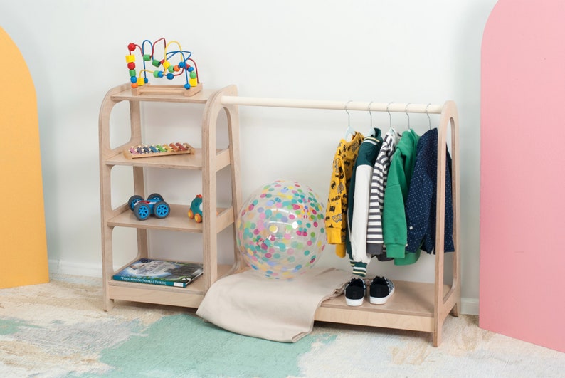 Op de afbeelding: Een houten kledingrek voor kinderen met een rek. Het rek heeft een hangende stang met kleding op hangers en een lager rek. Het rek heeft drie planken met speelgoed erop. Er ligt een kleurrijke bal op de vloer voor het rek.