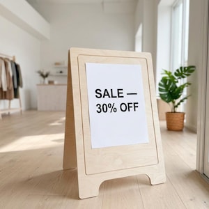 May include: Wooden A-frame sign with a white panel showing "SALE - 30% OFF" in black lettering. The sign is in a bright shop, with clothes racks and a potted plant visible in the background.