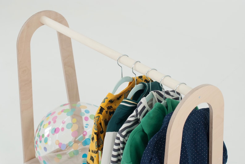 Op de afbeelding: Een houten kledingrek met een witte stang en een doorzichtige plastic bal met kleurrijk confetti. Het rek heeft een afgeronde bovenkant en houdt verschillende kleurrijke kinderkleding op hangers.