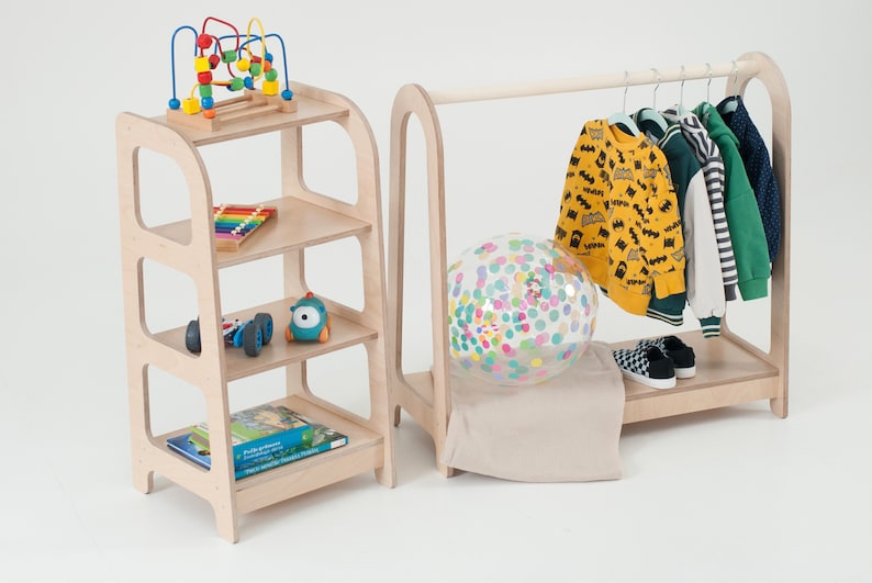 Op de afbeelding: Een houten kledingrek voor kinderen met een hangende stang en een lager plank. Het rek heeft een kleurrijke ballon en een paar kledingstukken die eraan hangen. Een houten plankeneenheid met drie planken is ook te zien, met speelgoed op de planken.