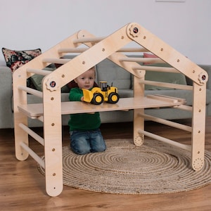 May include: A wooden climbing frame shaped like a house with a child playing inside. The frame has a ladder and a platform with a toy truck on it.