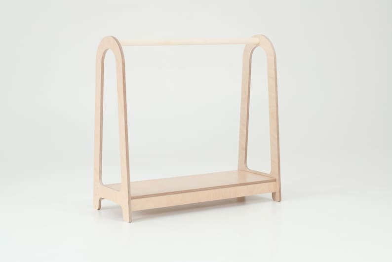 Op de afbeelding: Een kledingrek voor kinderen van licht hout met een afgeronde bovenste stang en een plank. Het rek is vrijstaand en heeft een eenvoudig ontwerp.